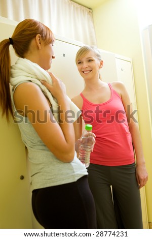 Two women stand in locker room and talking. One of them holding towel and bottle of water.