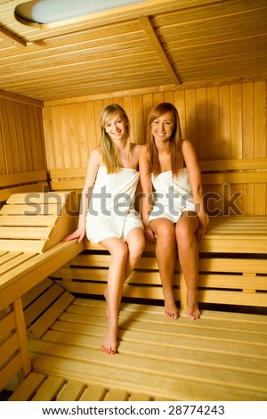 Two young women with towels sitting in sauna. They're looking at camera and smiling.