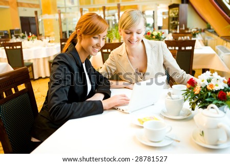 Two women sitting at table in restaurant. They're smiling and looking something on laptop.