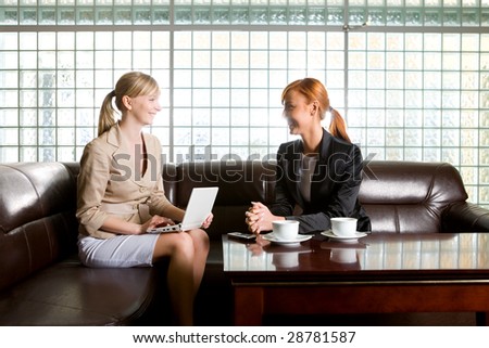 Two women sitting on couch and talking. One of them doing something on laptop.