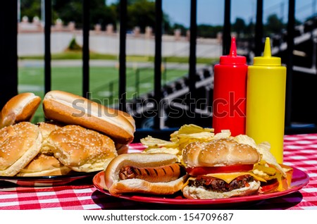 American football party with cheeseburger, hot dog, potato chips, ketchup and mustard bottles and buns.  Football field in background.