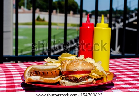 Tailgate party with cheeseburger, hot dog, potato chips and mustard and ketchup bottles.  Football field in background.