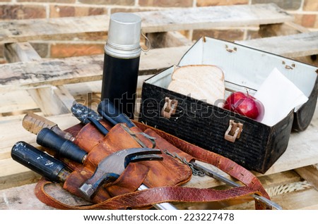 Lunch break at construction site with lunch pail, beverage container, apple, sandwich, napkin, tool belt, hammer, wrenches, screwdrivers on pallet with brick wall in background.