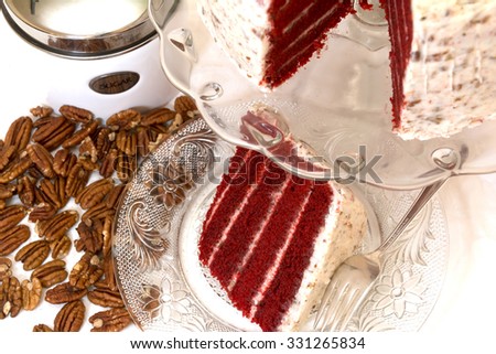 Slice of red velvet cake and pecans with sugar canister in background.  Slice is removed from whole cake which is in background.  Fork is on plate.