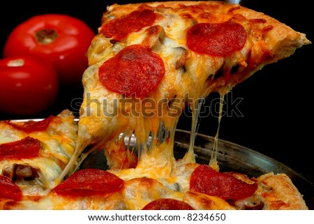Slice of pepperoni pizza being removed from whole pizza with tomatoes in background.  Isolated on black background.