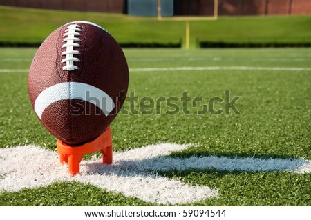 American football on tee on field with goal post in background.