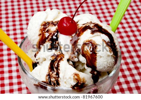 Vanilla ice cream and spoons with cherry and chocolate fudge topping on table with red gingham table cloth.
