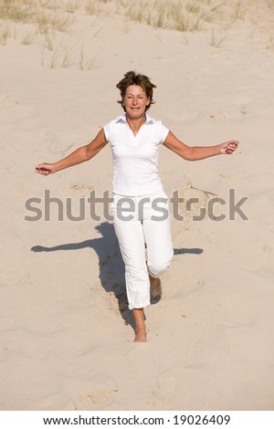 Active senior woman with white clothes is jumping through the sand.