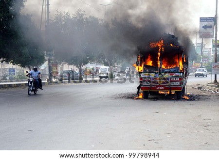 KARACHI, PAKISTAN - APR 11: Motorists pass through burning vehicle, which was set ablaze by unknown persons, at Mosamiyaat area during strike on the call of (JSQM) on April 11, 2012 in Karachi