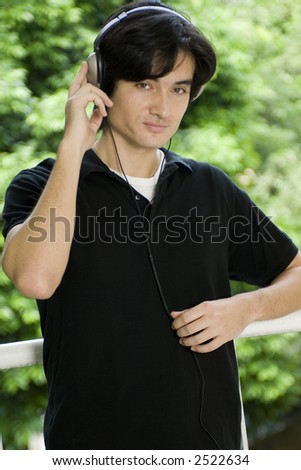 A young man standing on his balcony with trees behind listening to music on headphones