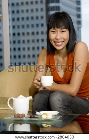 A young smiling asian woman drinking tea whilst sat at home on a sofa