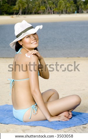 A young pretty asian woman wearing a sun hat sitting on tropical beach rubbing in suntan lotion