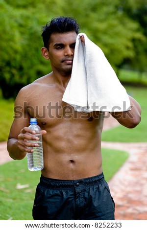 A well-built young man standing outside with no top with water bottle and towel