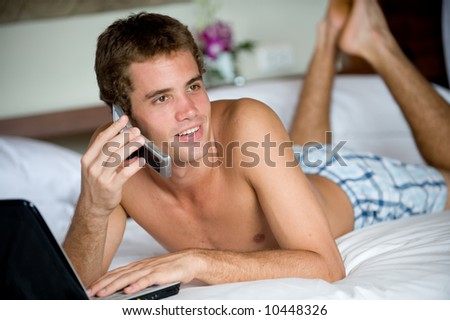 A young man using computer and phone whilst lying on bed