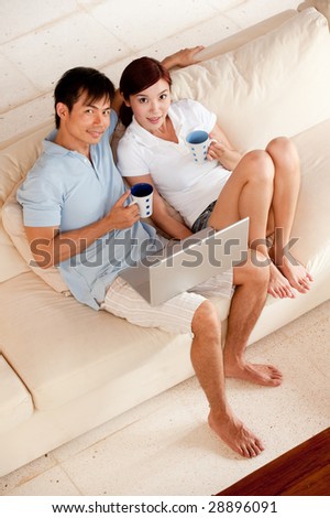 A young attractive Asian couple sitting at home on sofa with coffee and computer