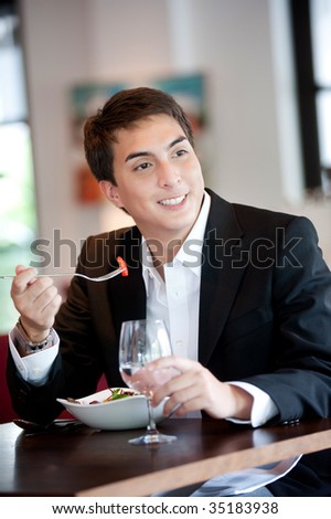 A young and attractive man uses his phone while eating a salad in an indoor restaurant