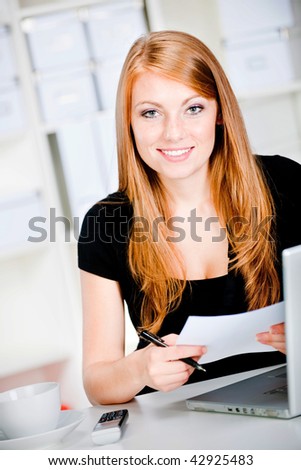 An attractive caucasian woman thinking with pen and paper in hand at the office, with a laptop, phone and a drink on the table