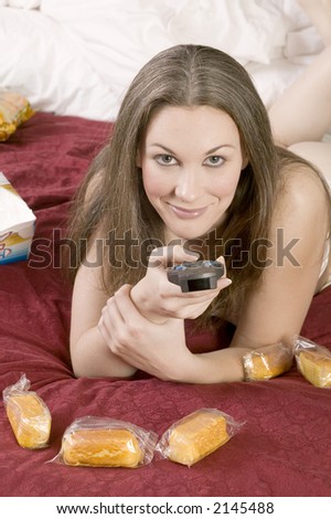 attractive young brunette woman in bed watching television and bing eating junk food