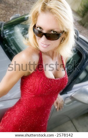 An attractive young blond woman in a red evening gown leans next to her sports car