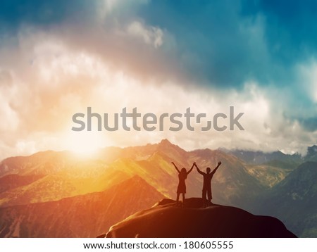 A happy couple standing together on the peak of a mountain with hands raised admiring breathtaking view at sunset