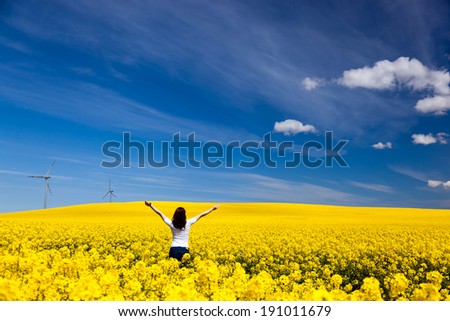 Happy young woman with hands raised on spring field of yellow flowers, rape. Blue sunny sky. Concepts of success, happiness, harmony, health, ecology