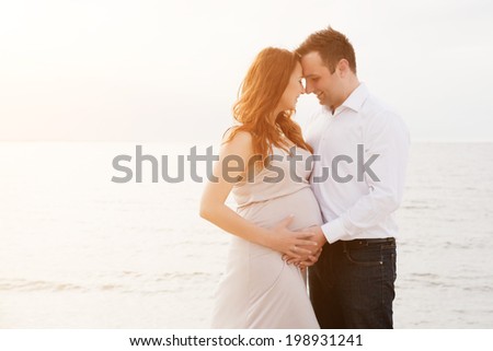 A pregnant beautiful woman with her husband on the beach smiling and touching her belly with love and care. Happy couple, relax by the calm sea in sunshine