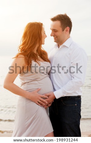 A pregnant beautiful woman with her husband on the beach smiling and touching her belly with love and care. Happy couple, relax by the calm sea in sunshine
