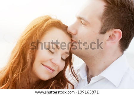 Young happy couple in love in a romantic moment of man kissing his wife in a forehead on the beach in summer sunshine.