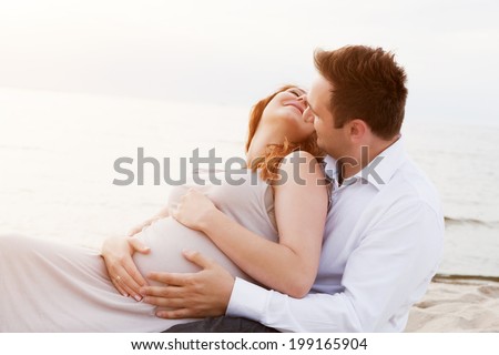 A pregnant beautiful woman with her husband on the beach having fun and touching her belly with love and care. Happy couple, relax by the calm sea in sunshine