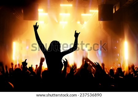 Concert, disco party. Woman silhouette with hands up in foreground and people having fun in night club