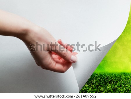 Hand pulling a bottom paper corner to uncover, reveal green landscape. Page curl, conceptual.