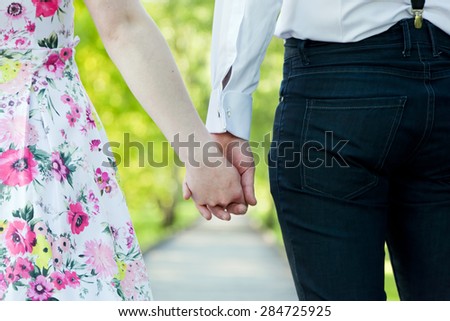 Young couple in love holding hands in summer park. Woman in dress and man wearing elegant shirt. View from the back. Date, fiance with fiancee, hand in hand concepts
