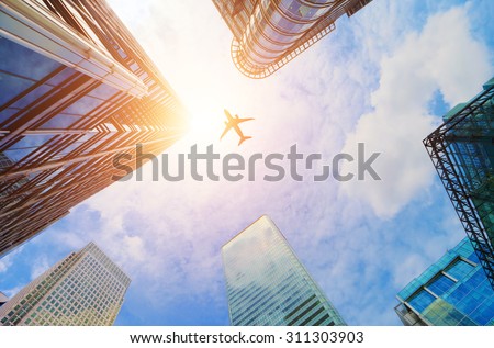 Airplane flying over business skyscrapers, high-rise buildings. Transport, transportation, travel. Sun light on blue sky.