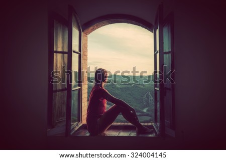 Young woman sitting in an open old window looking on the landscape of Tuscany, Italy. Conceptual romantic, dreaming, hope, travel.