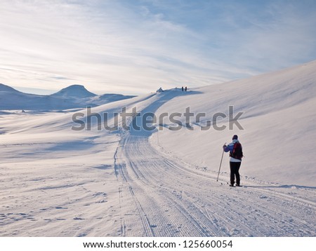 Female skier in a groomed ski track approaching a small hill with a mountain summit and light clouds in the background in the norwegian mountains