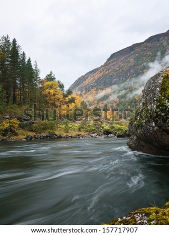 Smooth river passing through an autumn landscape with trees and cliffs and with low level clouds in the mountainside in the background