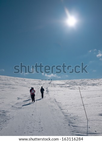 Family skiing uphill a mountainside in a groomed ski track following a group of people visible at the edge of a hill in the norwegian mountains at easter at a day with clear sky and sun in front