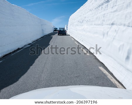 Road with cars at the snow road between Laerdal and Aurland in Norway early summer forming a narrow channel with dry asphalt and large snow edges on both sides