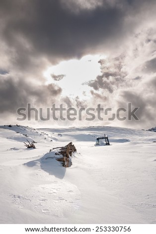 Snow covered wooden lodges together with a almost hidden tractor during an overcast day where sun nearly brakes through the clouds in the norwegian mountains at easter