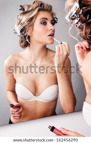 Girl in hair curlers applying lipstick while looking in a mirror.
