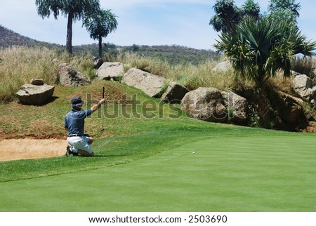 Middle age golfer sitting down next to the green, estimating the line for the putt.