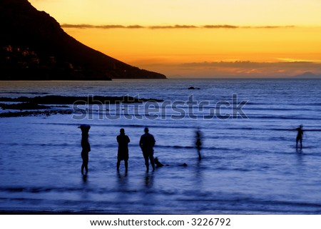 People standing in the ocean after sunset in Gordon\'s Bay, South Africa. Slow shutter speed results in some movement on people and the effect of foaming water.