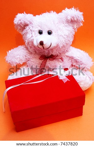 A pink fluffy bear sitting by a red box with a white bow