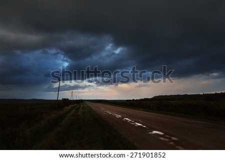 Road in the field and the dark dramatic rain clouds. Autumn in Latvia.