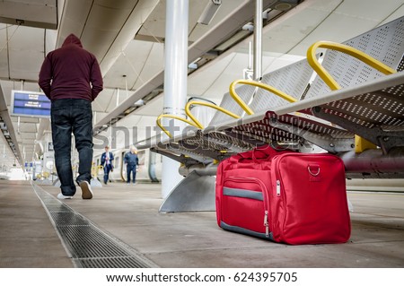Terrorism and public safety concept with an unattended bag left under chair on platform at train station or airport and man wearing a hoodie walking away from the suspicious item (possibly terrorist)
