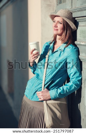 Beautiful young hipster woman in hat and blue jacket with cup of coffee outdoor