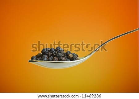 Metal spoon filled with black peppers over orange background