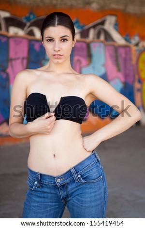Sexy brunette woman with jeans on graffiti wall take off her bustier.