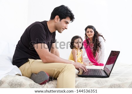 father working on the laptop with mother and daughter in the background, Indian family of three using laptop