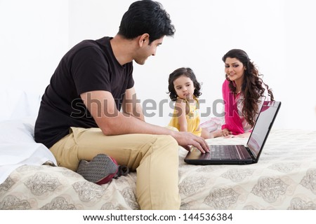 father working on the laptop with mother and daughter in the background, Indian family of three using laptop